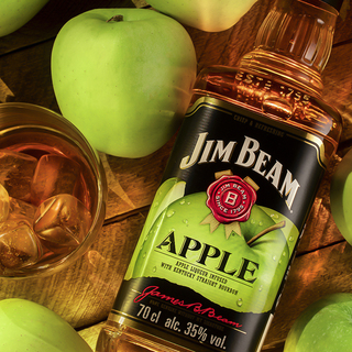 Still life with a bottle of Jim Beam Apple Bourbon , liqueur from Kentucky , with a glass of cold whiskey on ice on a wooden brown background of boards, surrounded by juicy green apples
