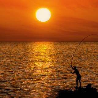 Silhouette Unrecognizable person of a fisherman on the background of round brilliant setting sun standing on a rock on the sea coast among water location horizontal