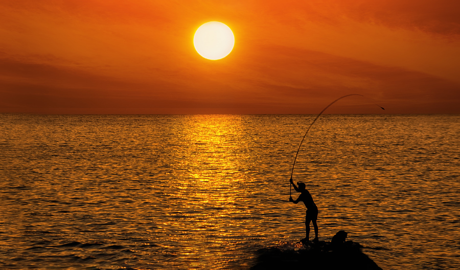 Silhouette Unrecognizable person of a fisherman on the background of round brilliant setting sun standing on a rock on the sea coast among water location horizontal