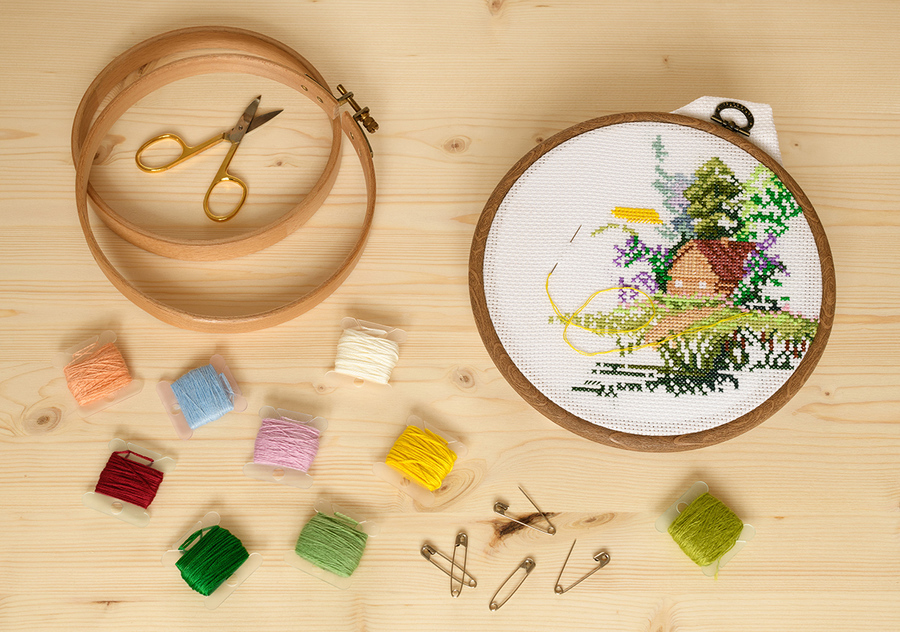 The embroidery house in round hoops and the accessories for needlework are on wooden table in indoor.