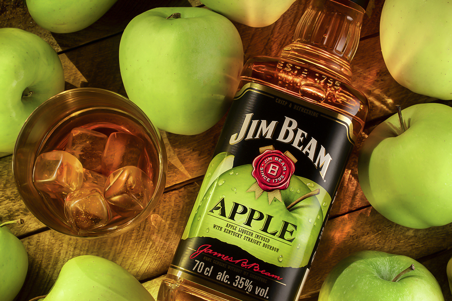 Still life with a bottle of Jim Beam Apple Bourbon , liqueur from Kentucky , with a glass of cold whiskey on ice on a wooden brown background of boards, surrounded by juicy green apples